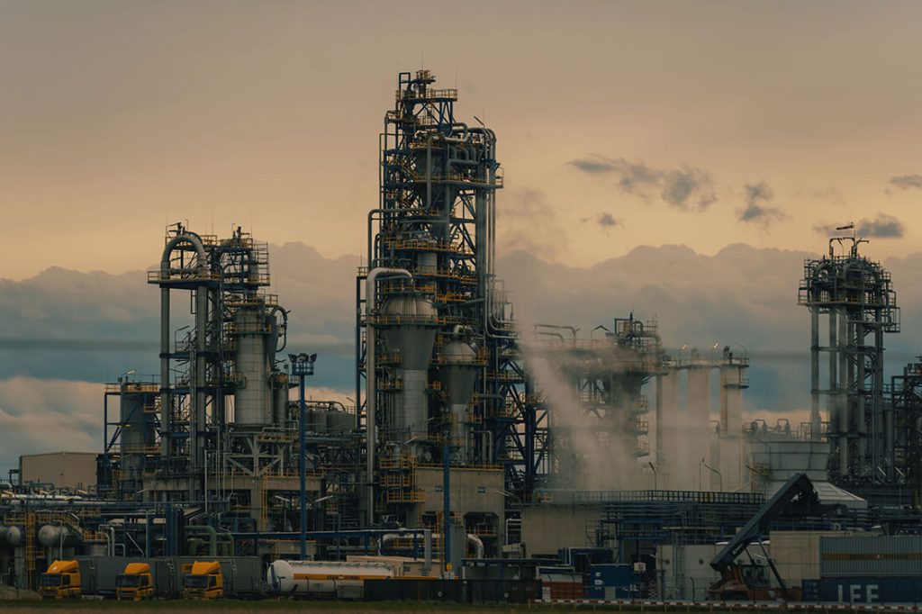 Large petrochemical refinery with distillation towers, tanks, pipes, and steam emissions under cloudy sunset sky.