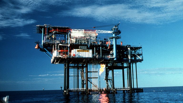 Offshore oil platform labeled Harriet A with cranes, pipes, and flare burning gas above calm ocean.
