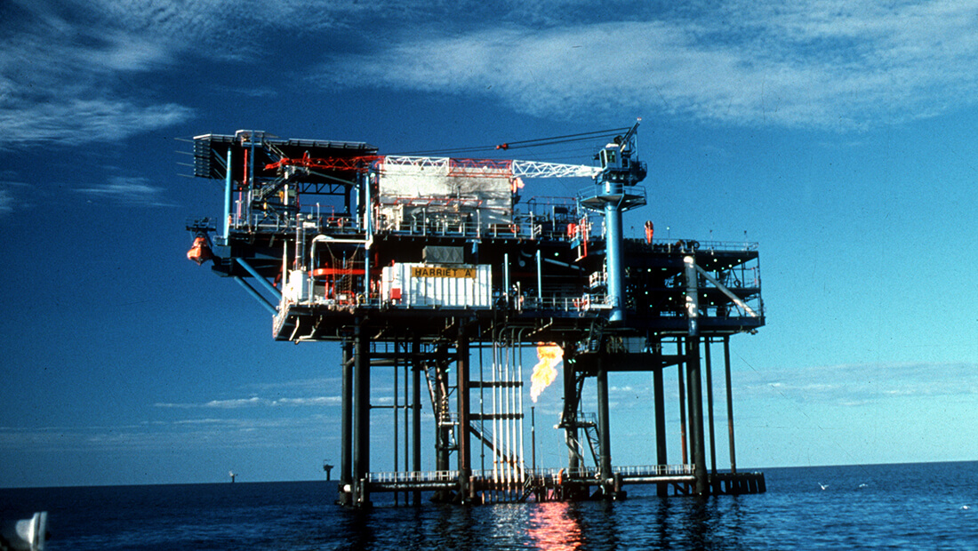 Offshore oil platform labeled Harriet A with cranes, pipes, and flare burning gas above calm ocean.