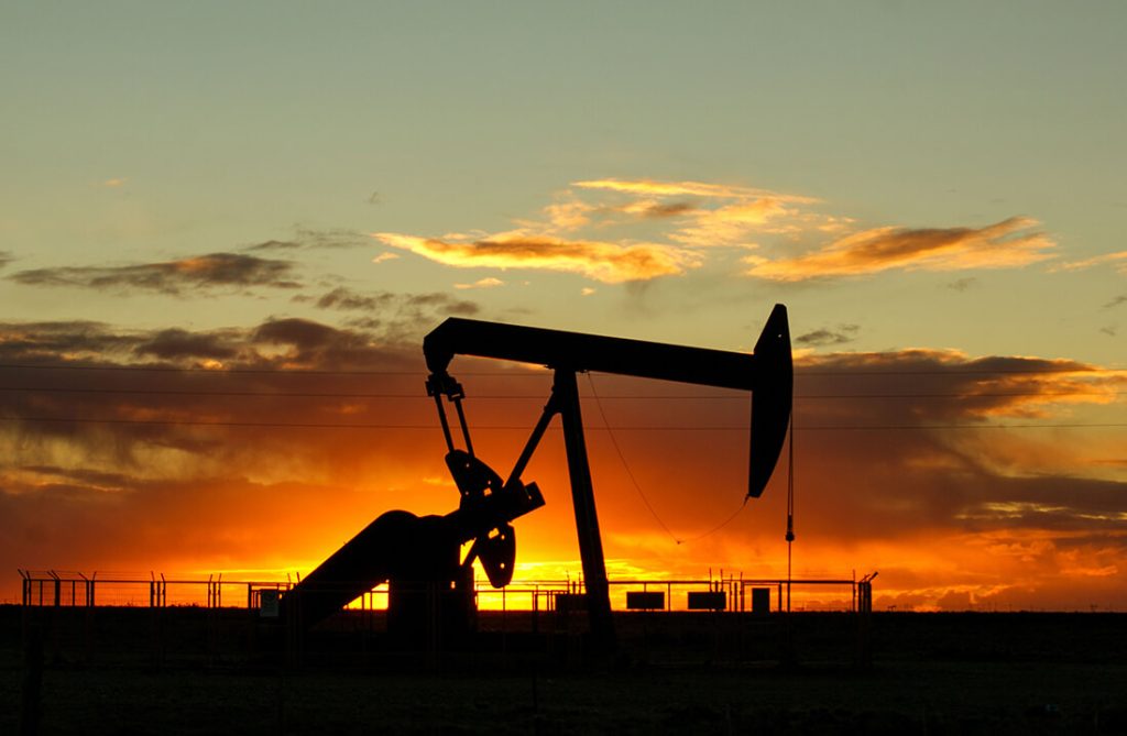 Silhouetted oil pumpjack against vivid sunset sky with orange, yellow, and blue gradient.
