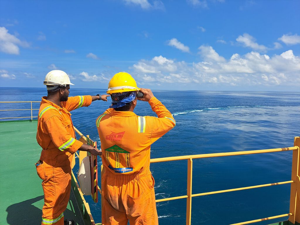 Two workers in orange gear on deck pointing at the ocean.
